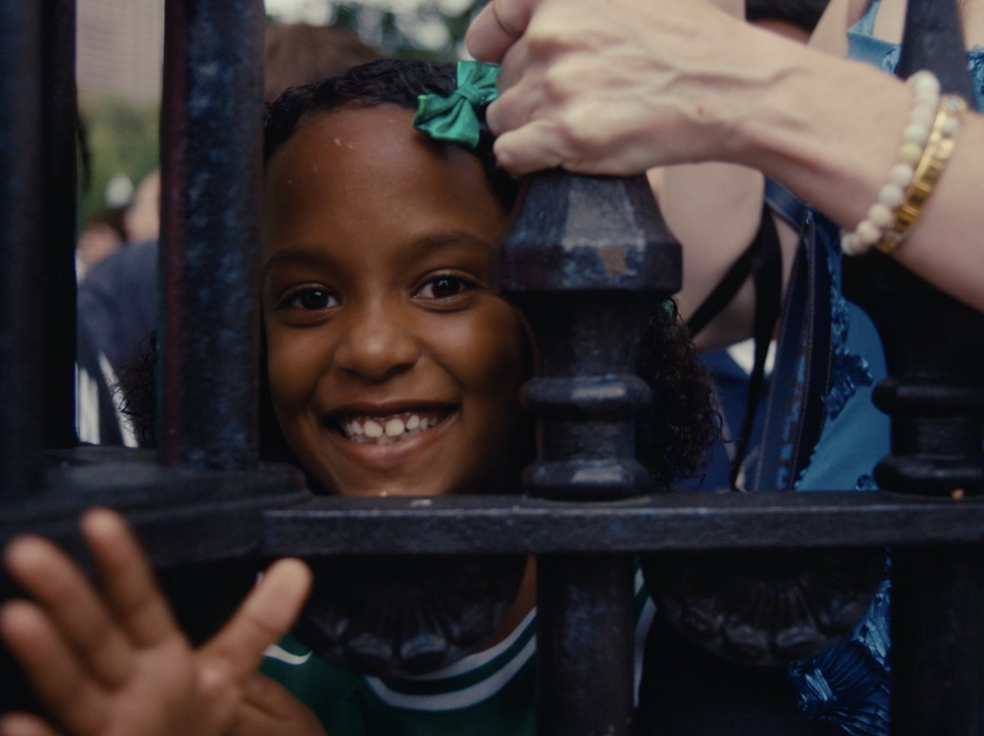 A young fan in a green bow peeking through a wrought-iron fence, waving