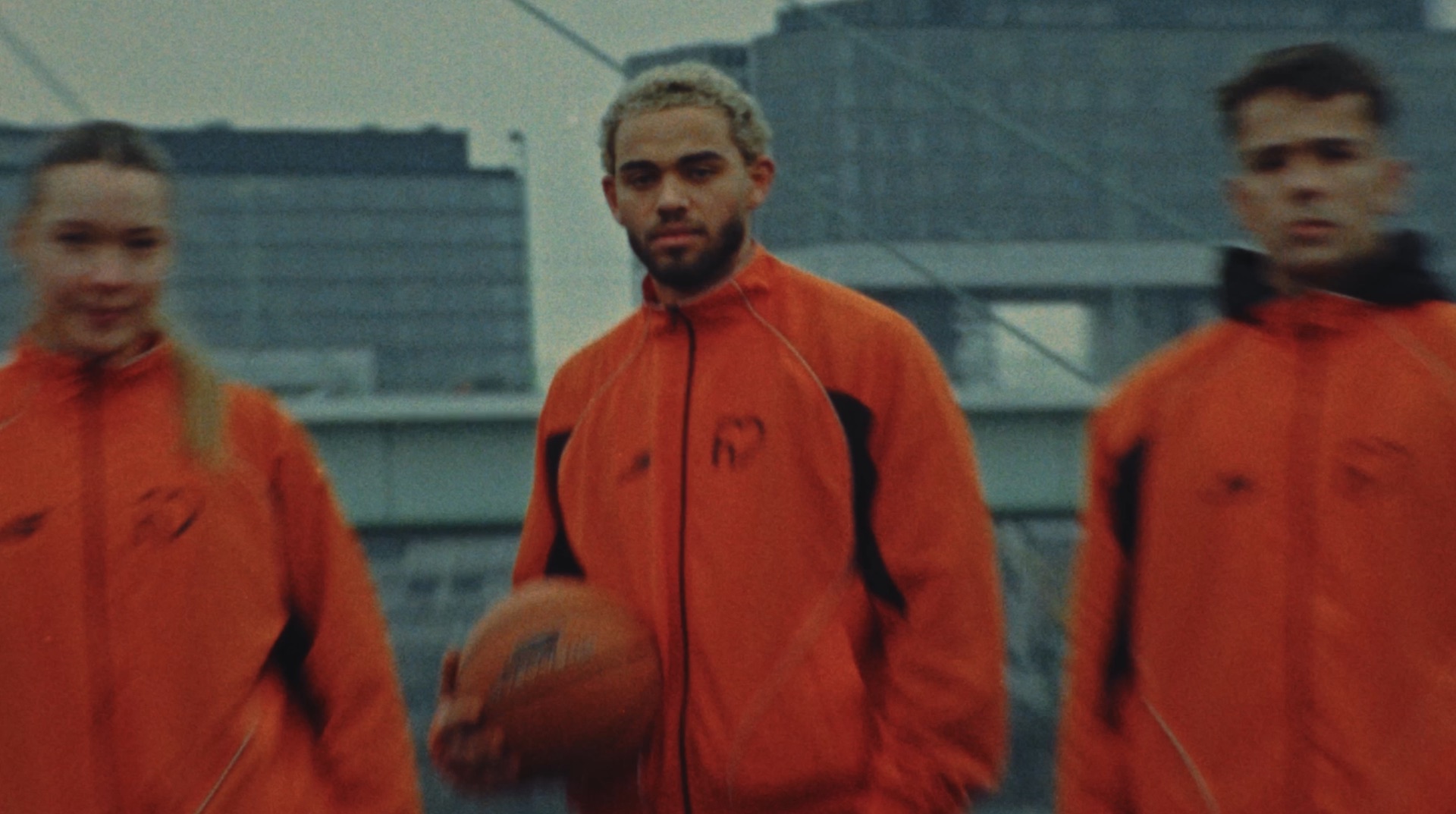 Three players in orange tracksuits with a heart-shaped logo, holding a basketball against a daylight cityscape