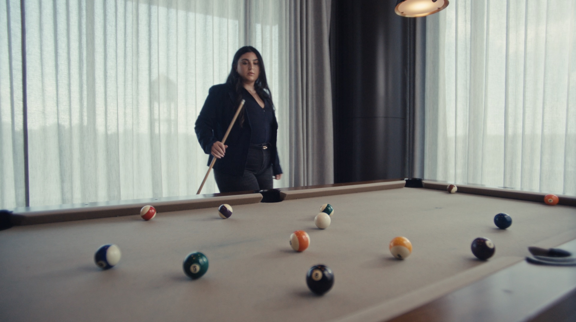 Woman lining up a shot at a pool table, curtained window behind her