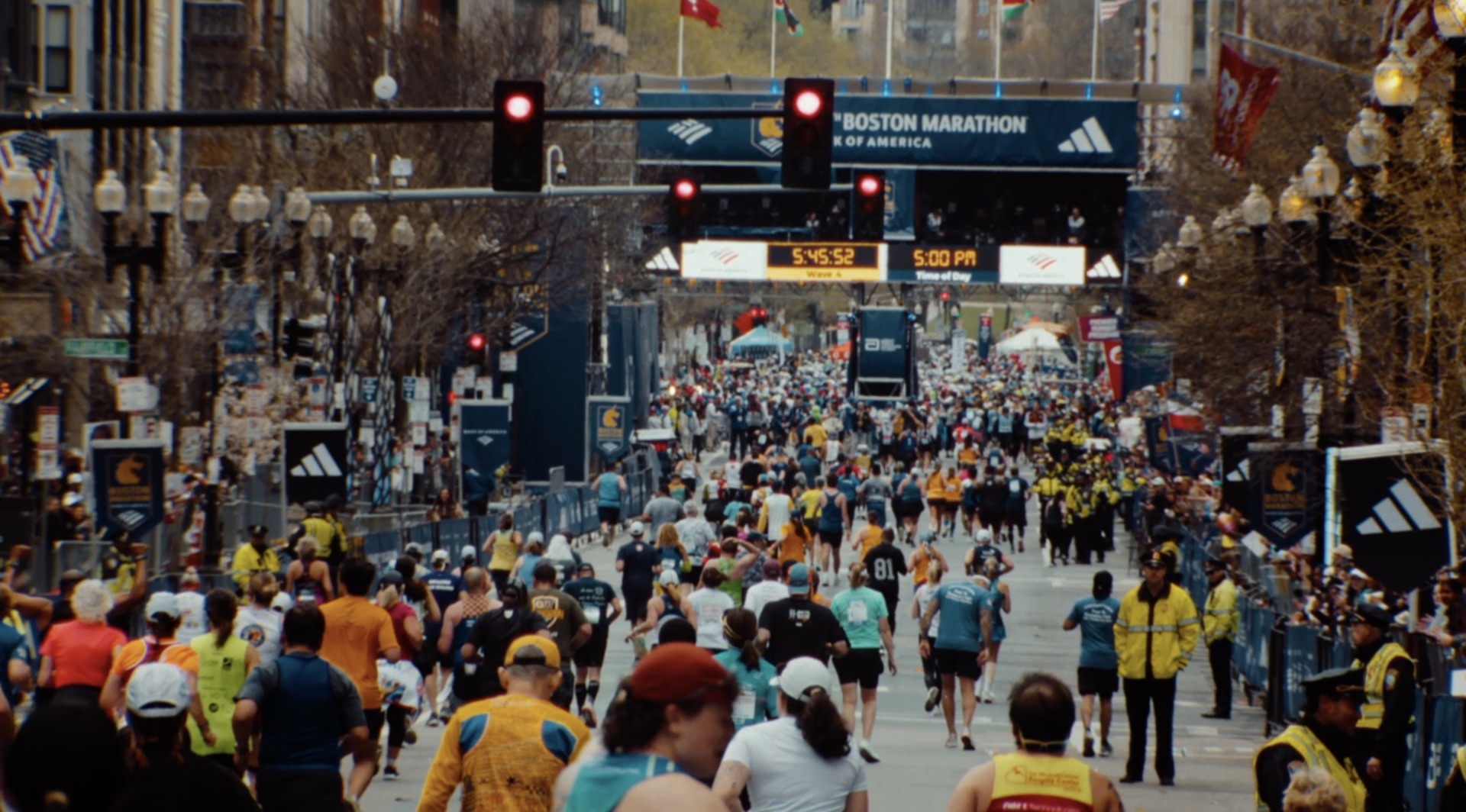 Wide shot of runners streaming past the Boston Marathon start banner, digital clock reading 5:45:52