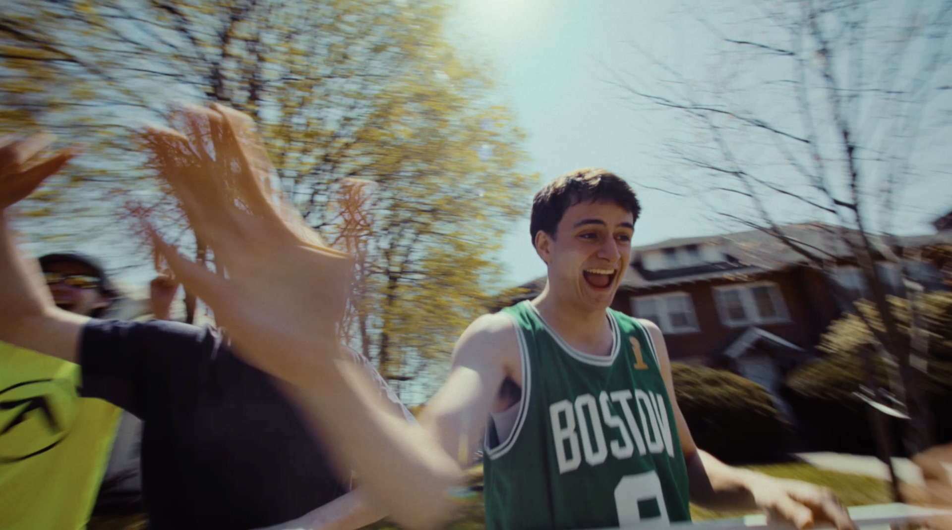 Spectator in a green Boston Celtics jersey cheering and high-fiving runners on a tree-lined street