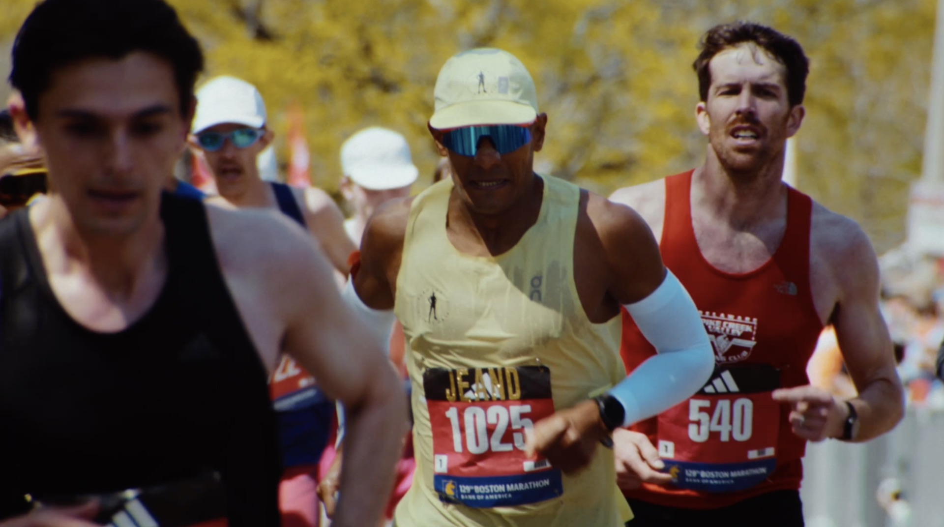 Lead pack of runners mid-race, bib numbers visible, sun and fall foliage behind them